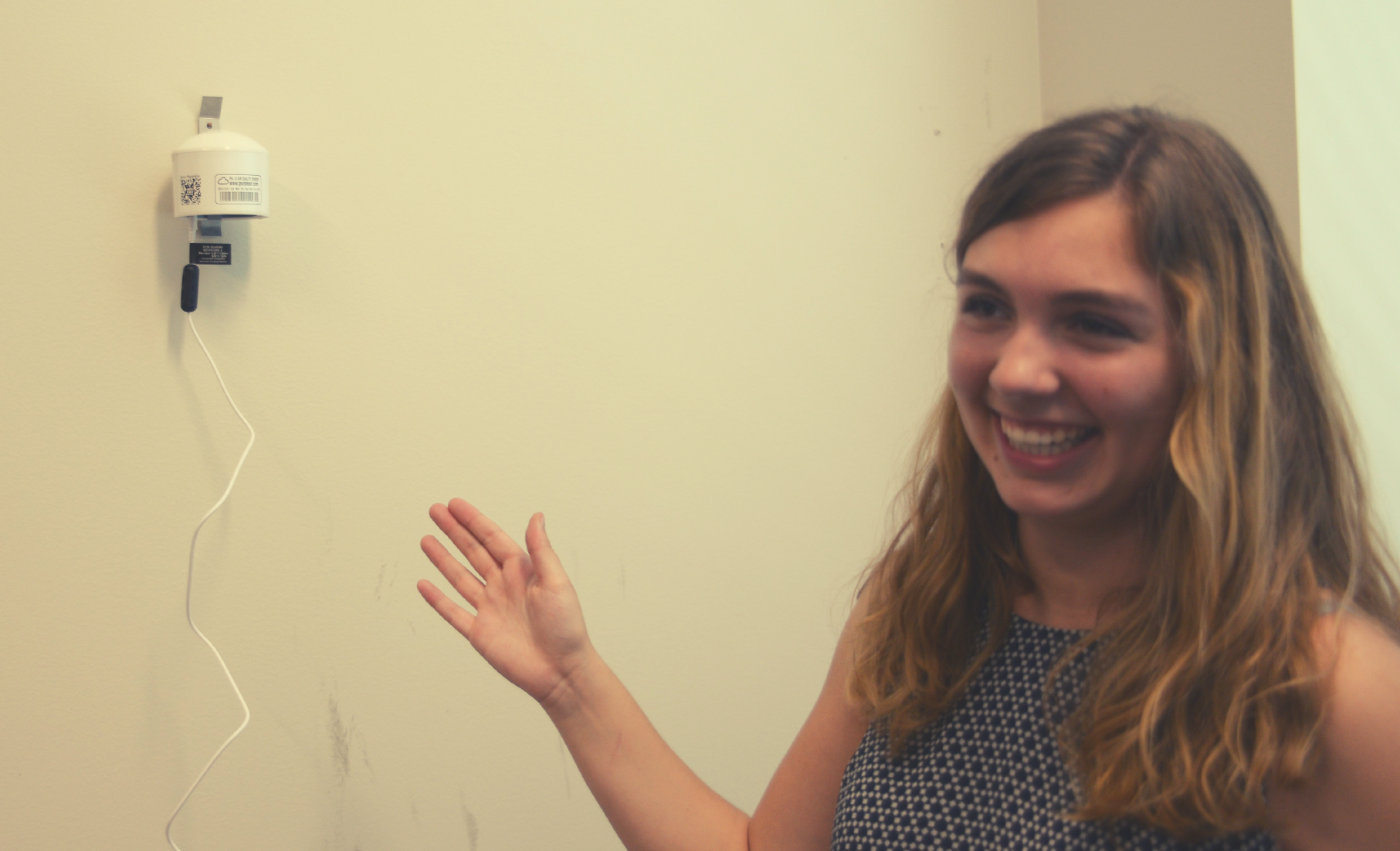 Image: student stands adjacent to wall-mounted PurpleAir instrument.