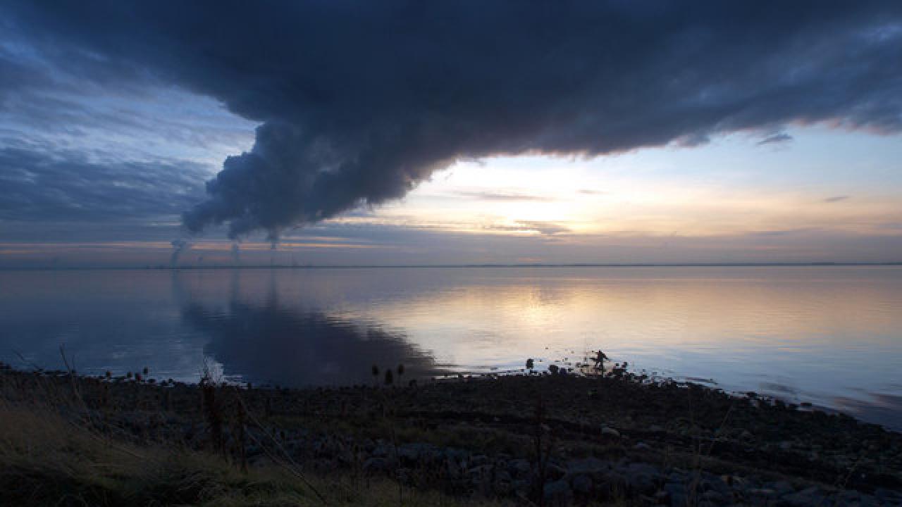 Image: a photo of a body of water with industrial stacks emitting large plumes of pollution.