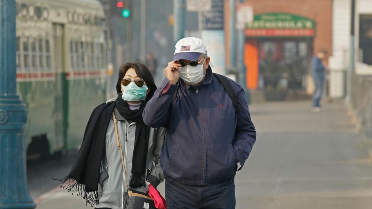 Image: two people stand outside a polluted downtown area wearing face masks.