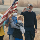 Image: people outside wear face masks and wave an American flag.