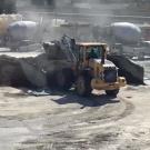 Image: a concrete plant featuring large vehicular equipment and visible dust plumes.